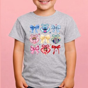 May include: A heather gray short-sleeve t-shirt featuring a central graphic print of various colorful bows and heart-shaped frames. Inside the heart frames are illustrations of Stitch, Angel, and Lilo, accented by small stars scattered across the design.