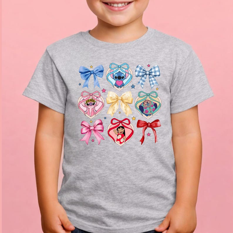 May include: A heather gray short-sleeve t-shirt featuring a central graphic print of various colorful bows and heart-shaped frames. Inside the heart frames are illustrations of Stitch, Angel, and Lilo, accented by small stars scattered across the design.