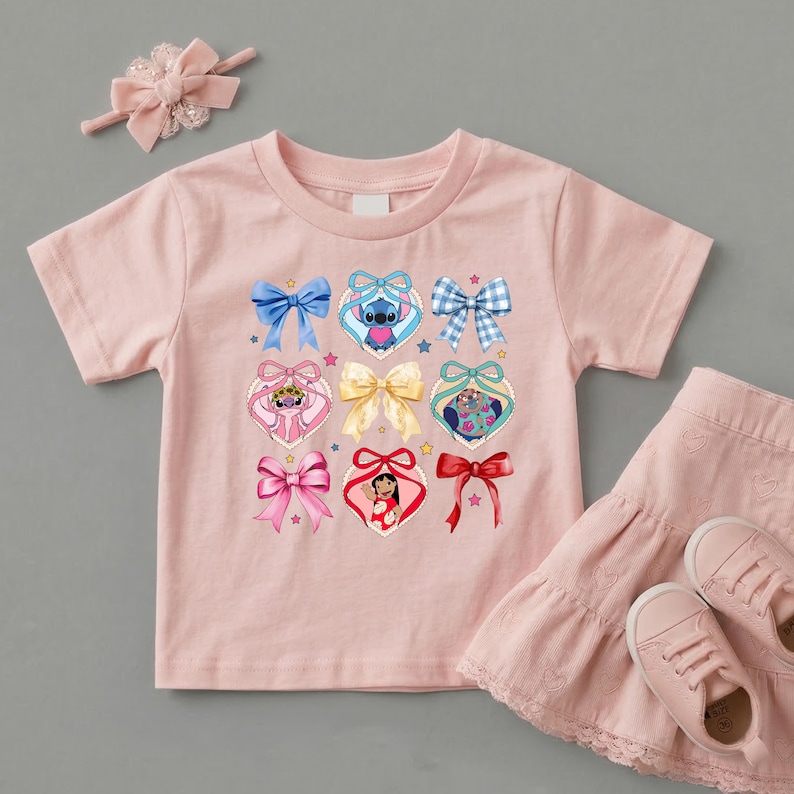 May include: A light pink short-sleeve t-shirt featuring a graphic print of various bows and heart-shaped frames containing cartoon characters. The set includes a matching light pink hair bow with lace trim and a light pink skirt with sneakers.
