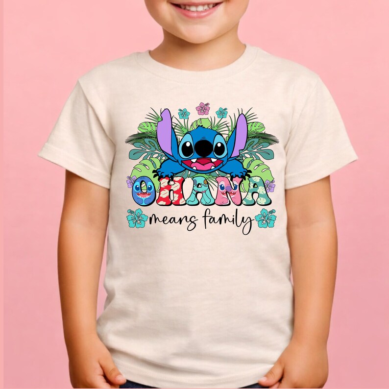 May include: A cream-colored short-sleeve t-shirt featuring a graphic of the blue character Stitch surrounded by green tropical leaves and colorful hibiscus flowers. The text "OHANA" is printed in large, patterned letters with "means family" below.