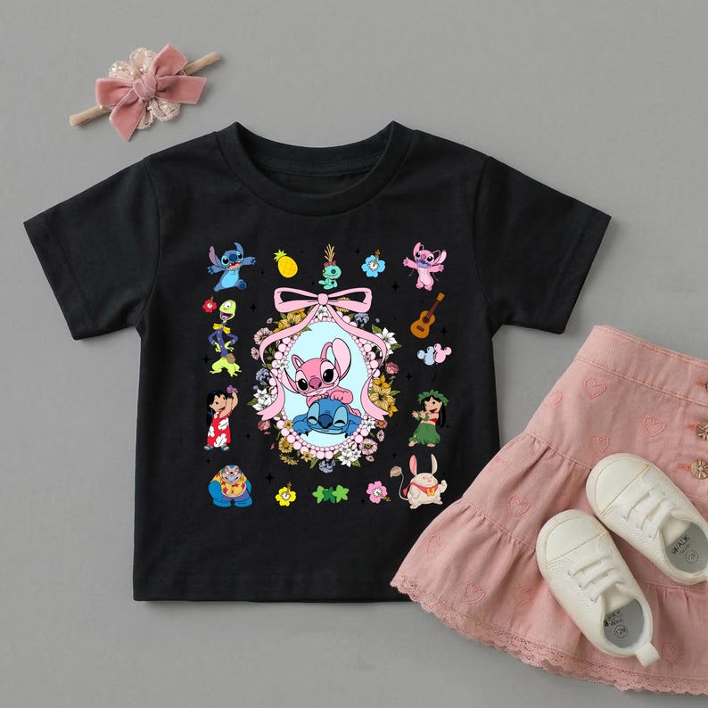 May include: A black short-sleeved t-shirt featuring a central oval frame decorated with flowers and a pink ribbon, containing illustrations of Stitch and Angel. Surrounding the frame are various small character graphics, including Lilo, Scrump, and tropical icons.
