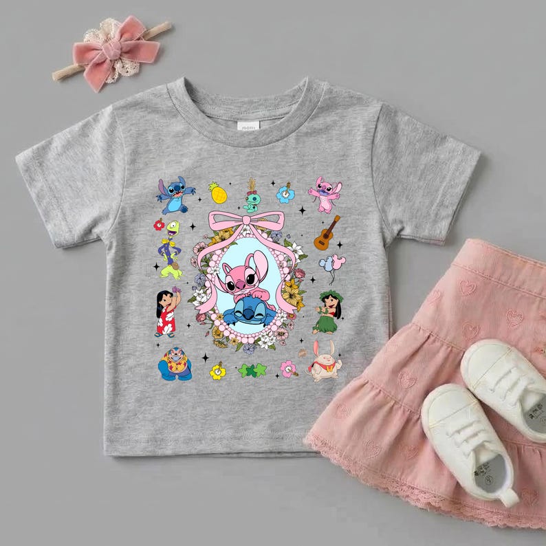 May include: A heather gray short-sleeve t-shirt featuring a central oval frame with a pink and blue alien character, surrounded by a floral wreath and a pink bow. Smaller illustrations of various cartoon characters and items surround the central frame.