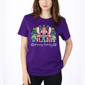 May include: A purple short-sleeved t-shirt featuring a graphic of a pink alien character surrounded by green tropical leaves and hibiscus flowers. The text reads OHANA in colorful patterned letters, followed by the script text means family.