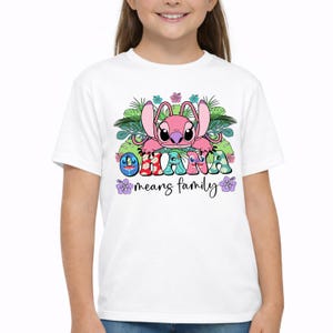 May include: A white short-sleeved t-shirt featuring a graphic of a pink alien character surrounded by green tropical leaves and colorful hibiscus flowers. The text OHANA is printed in patterned letters, with the phrase means family written below it.