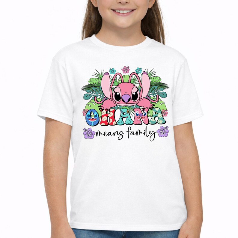 May include: A white short-sleeved t-shirt featuring a graphic of a pink alien character surrounded by green tropical leaves and colorful hibiscus flowers. The text OHANA is printed in patterned letters, with the phrase means family written below it.