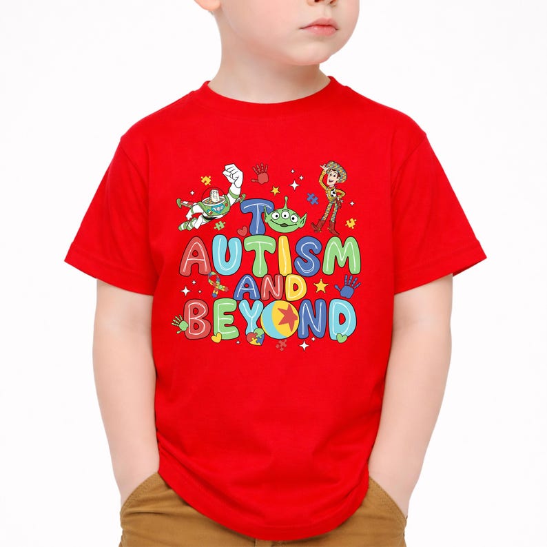 Op de afbeelding: Een rood T-shirt met de woorden "AUTISM AND BEYOND" in kleurrijke letters. Het shirt heeft cartoonafbeeldingen van personages. Het shirt wordt door een kind gedragen.