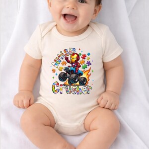 Puede incluir: Un body de beb&eacute; color crema con un gr&aacute;fico colorido que presenta un personaje de dibujos animados de Iron Man conduciendo un monster truck. El texto en el body dice "Autism Crusher" en una fuente de arco&iacute;ris, rodeado de piezas de rompecabezas.