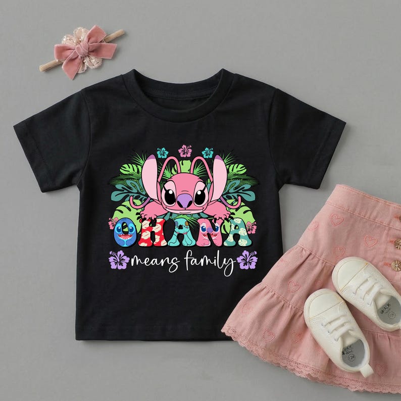 May include: A black short-sleeved t-shirt featuring a pink character graphic, tropical leaves, and hibiscus flowers. The text reads "OHANA means family" in colorful, patterned letters. A pink hair bow and a pink skirt with white sneakers are included.