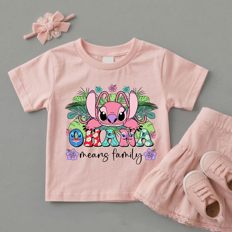 May include: A light pink short-sleeved t-shirt featuring a graphic of a pink alien character surrounded by green tropical leaves and hibiscus flowers. The text "OHANA means family" is printed in colorful, patterned letters below the character.