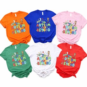 Op de afbeelding: Zes shirts met korte mouwen in oranje, blauw, roze, groen, wit en rood. Elk shirt toont de tekst "AUTISM AND BEYOND" met stripfiguren. De shirts zijn gemaakt van een zacht materiaal.
