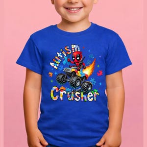 Puede incluir: Una camiseta azul con un personaje de dibujos animados de Deadpool conduciendo un cami&oacute;n monstruo con llamas. El texto "Autism Crusher" se muestra en letras de rompecabezas de colores. La camiseta la lleva un ni&ntilde;o.