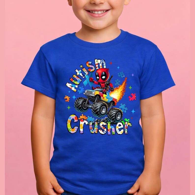 Puede incluir: Una camiseta azul con un personaje de dibujos animados de Deadpool conduciendo un cami&oacute;n monstruo con llamas. El texto "Autism Crusher" se muestra en letras de rompecabezas de colores. La camiseta la lleva un ni&ntilde;o.