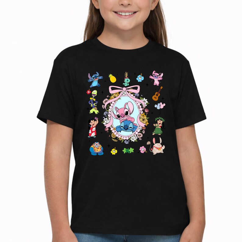 May include: A black short-sleeved t-shirt featuring a central oval frame decorated with flowers and a pink ribbon, containing a pink and blue alien character. Surrounding the frame are various small colorful cartoon characters and icons.