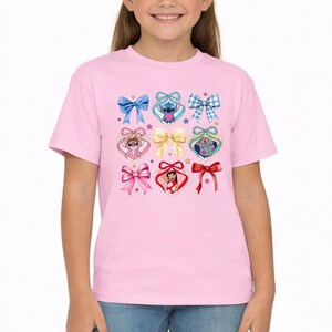 May include: A light pink short-sleeved t-shirt featuring a graphic print of various bows and heart-shaped frames. The design includes illustrations of Stitch and Lilo from Lilo and Stitch, interspersed with solid, patterned, and ribbon-style bows.