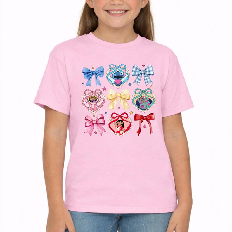 May include: A light pink short-sleeved t-shirt featuring a graphic print of various bows and heart-shaped frames. The design includes illustrations of Stitch and Lilo from Lilo and Stitch, interspersed with solid, patterned, and ribbon-style bows.