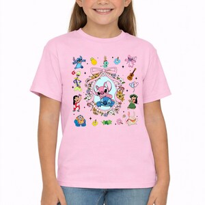 May include: A light pink short-sleeve t-shirt featuring a central graphic of two cartoon characters inside a floral oval frame with a bow. The design includes various small icons like a pineapple, guitar, and additional character illustrations.