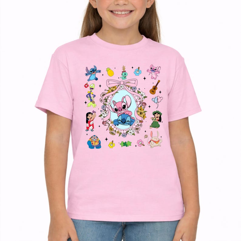 May include: A light pink short-sleeve t-shirt featuring a central graphic of two cartoon characters inside a floral oval frame with a bow. The design includes various small icons like a pineapple, guitar, and additional character illustrations.