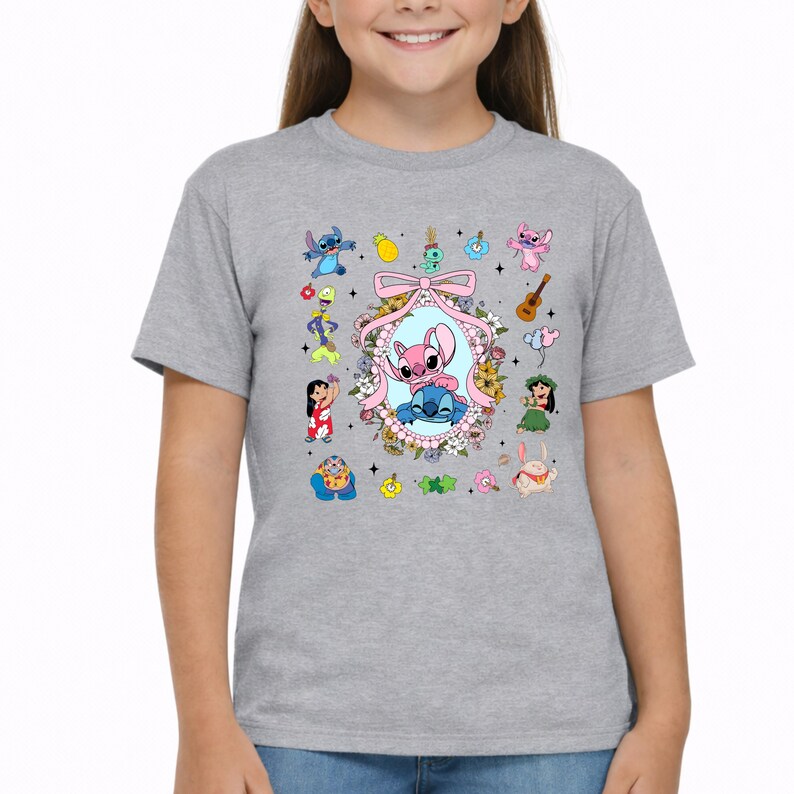 May include: A heather gray short-sleeve t-shirt featuring a central oval frame decorated with flowers and a pink bow, containing illustrations of Stitch and Angel. Surrounding the frame are various small character icons, including Lilo, a guitar, and tropical elements.
