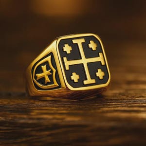 Puede incluir: Un anillo dorado con un centro cuadrado negro con un diseño de cruz. El anillo tiene un diseño de escudo en el lateral con una cruz. El anillo está sobre una superficie de madera.