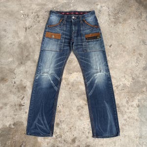 Op de afbeelding: Blauwe jeans van denim met bruine su&egrave;de-achtige zakaccenten en ritssluitingsdetails. De jeans hebben een vervaagde wassing en een klassiek ontwerp met vijf zakken. De jeans liggen plat op een betonnen oppervlak.