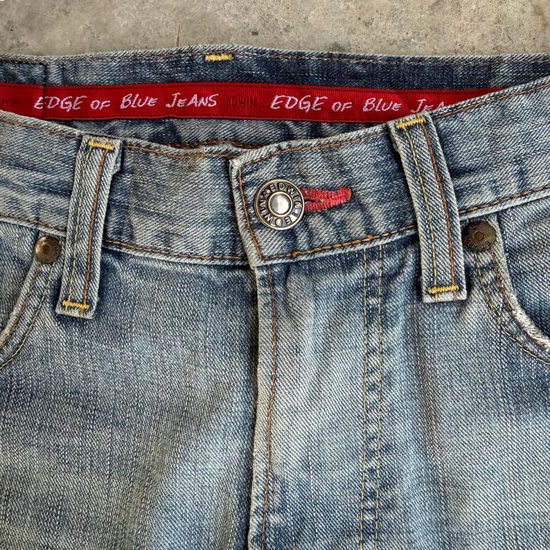 May include: Close-up of light blue denim jeans. The jeans feature a button closure, belt loops, and a red waistband with the text "EDGE OF BLUE JEANS". The stitching is visible, and the jeans appear worn.