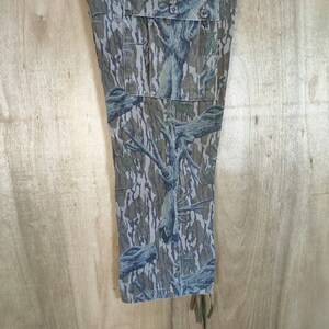 May include: Close-up of camouflage trousers with a tree bark pattern in green, brown, and grey. The trousers have pockets and button fastenings. The fabric has a textured appearance, and the design is detailed and realistic.