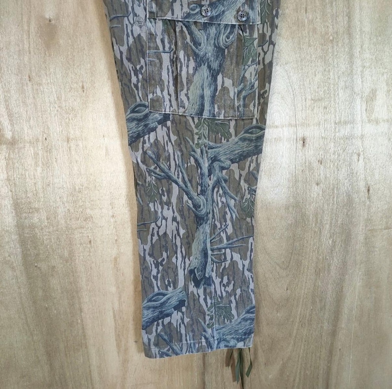 May include: Close-up of camouflage trousers with a tree bark pattern in green, brown, and grey. The trousers have pockets and button fastenings. The fabric has a textured appearance, and the design is detailed and realistic.