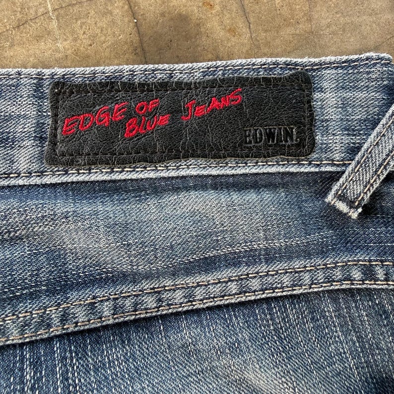 以下が含まれることがあります： 青いデニムジーンズのクローズアップ。黒い革のパッチには、赤い文字で「EDGE OF BLUE JEANS」とブランド名「EDWIN」が書かれています。ジーンズにはステッチが見えます。