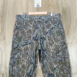 May include: Camouflage cargo trousers with a tree bark pattern in shades of brown, green, and grey. The trousers have multiple pockets and a button closure. The trousers are hanging on a wooden hanger.