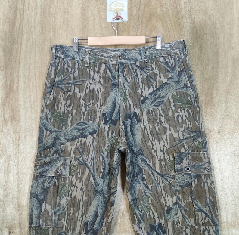 May include: Camouflage cargo trousers with a tree bark pattern in shades of brown, green, and grey. The trousers have multiple pockets and a button closure. The trousers are hanging on a wooden hanger.