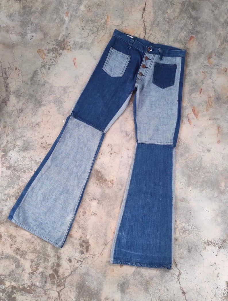May include: Two-tone denim jeans with a button fly and two front pockets. The jeans feature a patchwork design with light and dark blue denim panels. The legs flare out from the knee, creating a bell-bottom style.