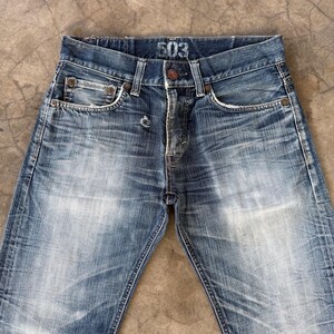 May include: Faded blue denim jeans with a button closure and five-pocket design. The jeans have a worn appearance with visible fading and distressing. The number "503" is printed on the waistband. A classic, casual style.