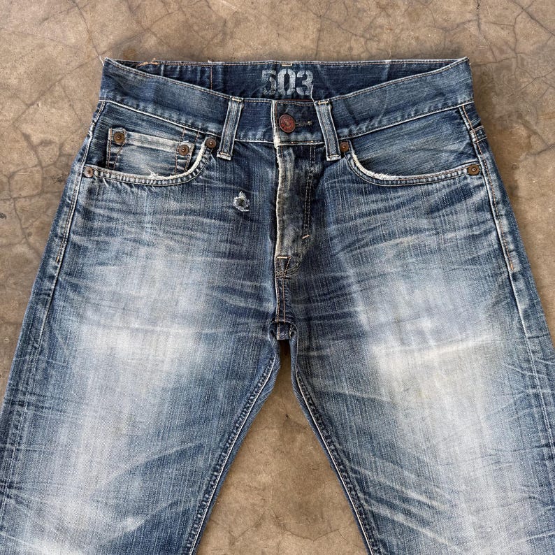 May include: Faded blue denim jeans with a button closure and five-pocket design. The jeans have a worn appearance with visible fading and distressing. The number "503" is printed on the waistband. A classic, casual style.