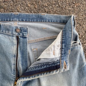 May include: Close-up of light blue denim jeans with a partially unzipped brass zipper. The jeans have a button closure and visible stitching. The fabric shows signs of wear and fading, suggesting a vintage or well-worn appearance.
