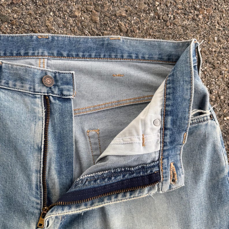 May include: Close-up of light blue denim jeans with a partially unzipped brass zipper. The jeans have a button closure and visible stitching. The fabric shows signs of wear and fading, suggesting a vintage or well-worn appearance.