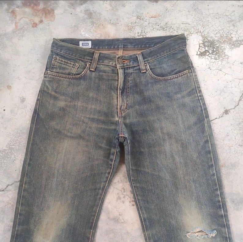 May include: Faded blue denim jeans with a distressed look. Features a classic five-pocket design, a button and zipper closure, and a small brand tag. The jeans have a worn appearance with a ripped knee.