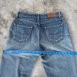 May include: Light blue denim jeans with a faded wash. The jeans have a classic five-pocket design, a brown leather label that says "SOMETHING", and a red stitch detail. A blue measuring tape shows a measurement of 22 inches.