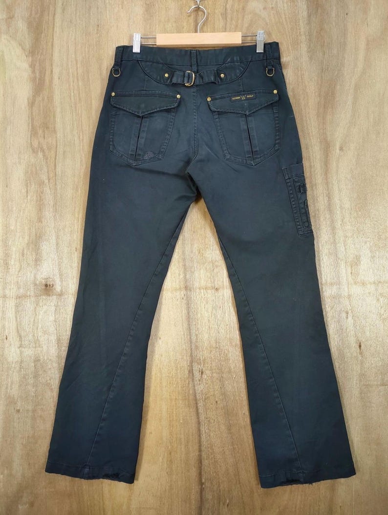 Pu&ograve; includere: Pantaloni blu navy scuro con tasche multiple e cintura. I pantaloni hanno un design a gamba svasata e dettagli dorati. Il retro presenta due tasche con patta e una cintura con fibbia. I pantaloni sono appesi a una gruccia di legno.