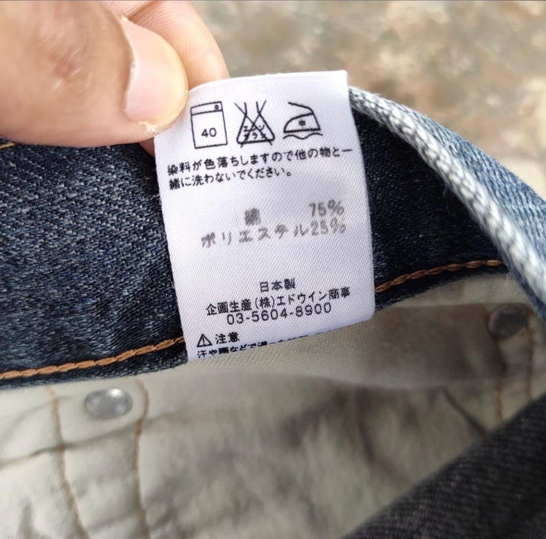 Puede incluir: Primer plano de una etiqueta blanca con texto y s&iacute;mbolos japoneses, adherida a una tela vaquera azul. La etiqueta incluye instrucciones de lavado, porcentajes de composici&oacute;n de la tela y un n&uacute;mero de tel&eacute;fono.