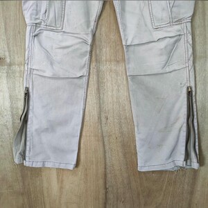 May include: Light gray cargo pants with multiple pockets and zippers on the lower legs. The pants have a slightly worn appearance and are made of a durable fabric. The design includes reinforced stitching and a classic cargo style.