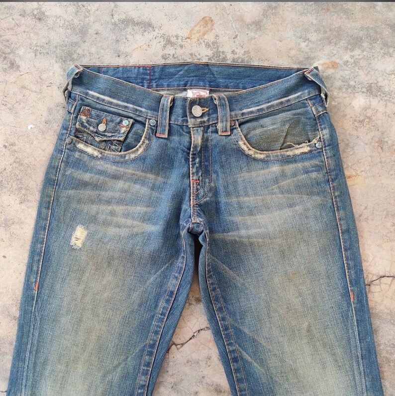 May include: Faded blue denim jeans with a distressed appearance. They have a button closure, front pockets, and decorative stitching. The jeans have a worn look with a small tear on the left leg, and a light wash.