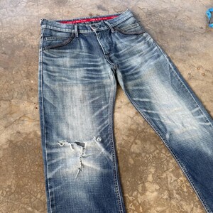 以下が含まれることがあります： 色あせた青色のデニムジーンズ。膝に破れがあり、ダメージ加工が施されています。ジーンズは、クラシックな5ポケットデザイン、赤いウエストバンドのディテール、ボタンとジッパーの留め具を備えています。デニムは使用感があります。