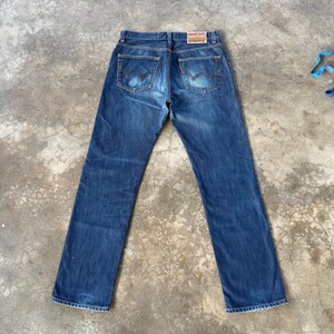 May include: Blue denim jeans with a classic five-pocket design. The jeans feature a faded wash and a red tag on the back pocket. The back pockets have a distinctive curved stitching pattern.