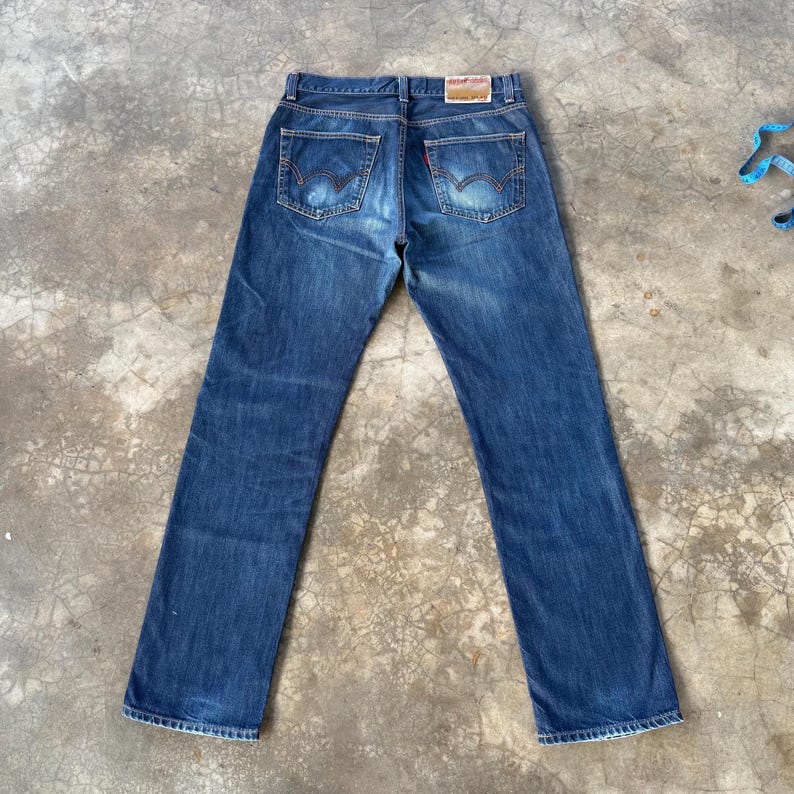 May include: Blue denim jeans with a classic five-pocket design. The jeans feature a faded wash and a red tag on the back pocket. The back pockets have a distinctive curved stitching pattern.