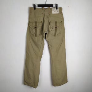 May include: Khaki-colored cargo pants with multiple pockets and a straight leg design. The pants are hanging on a black hanger against a neutral background. The back of the pants is visible, showcasing the pocket details.