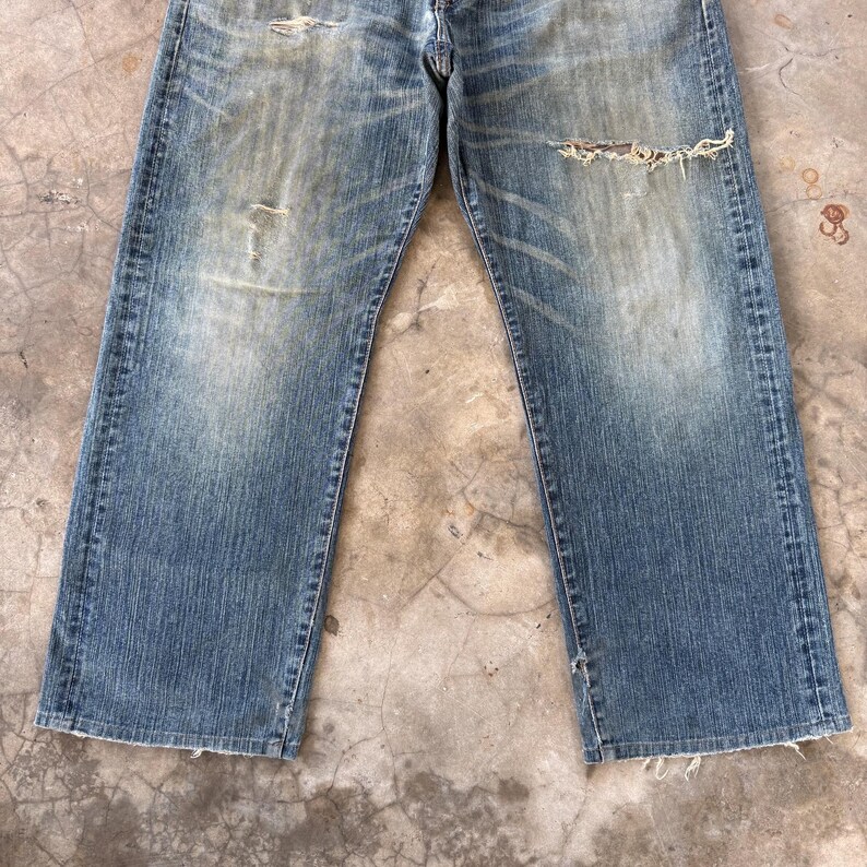 Puede incluir: Vaqueros de mezclilla azul descolorido con detalles desgastados, incluyendo rasgaduras y parches desgastados. Los vaqueros tienen un dise&ntilde;o cl&aacute;sico de cinco bolsillos y un corte de pierna recta. El denim tiene un lavado vintage, d&aacute;ndole un aspecto desgastado.