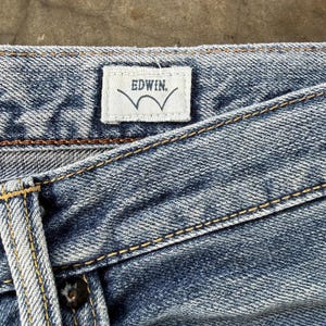 Pu&ograve; includere: Primo piano di jeans di denim blu con cuciture visibili e un'etichetta bianca. L'etichetta riporta "EDWIN" in un carattere sans-serif sopra un disegno a onda stilizzato.