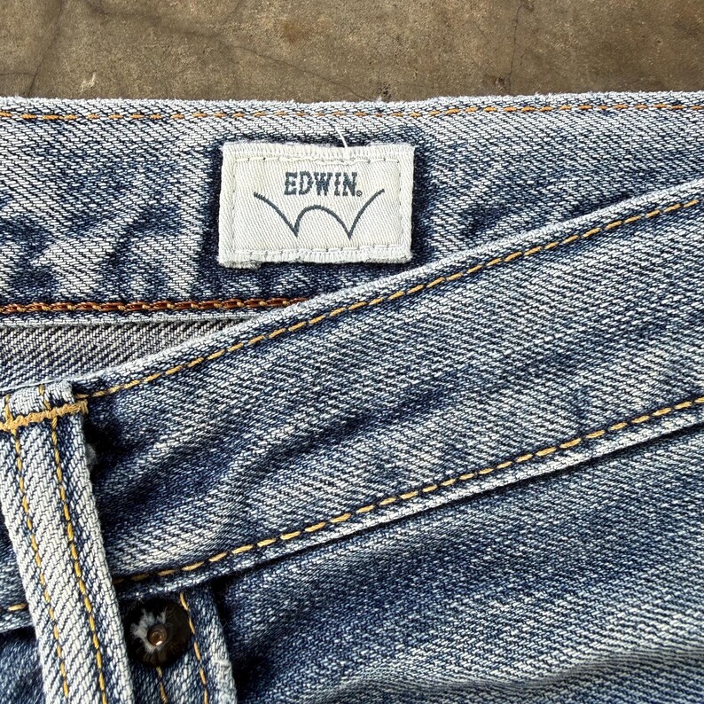 Pu&ograve; includere: Primo piano di jeans di denim blu con cuciture visibili e un'etichetta bianca. L'etichetta riporta "EDWIN" in un carattere sans-serif sopra un disegno a onda stilizzato.