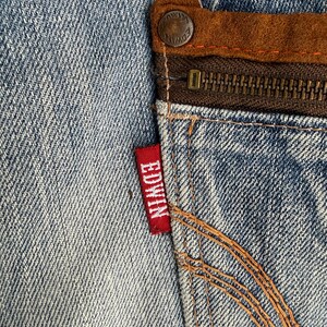 May include: Close-up of a section of blue denim jeans. The image highlights the details of the fabric, stitching, and a red "EDWIN" tag. A brown leather patch and a zipper are also visible.