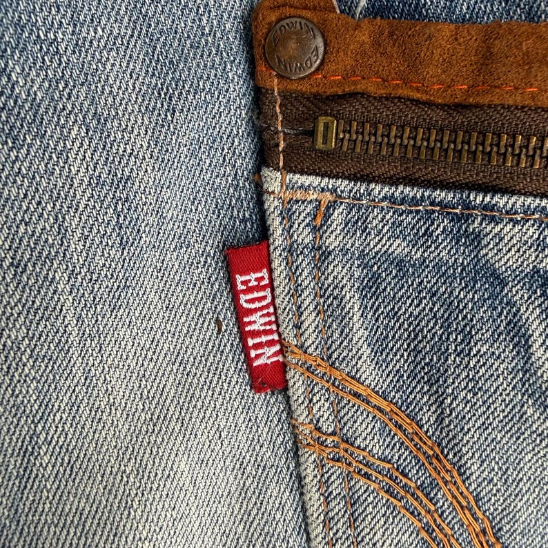 May include: Close-up of a section of blue denim jeans. The image highlights the details of the fabric, stitching, and a red "EDWIN" tag. A brown leather patch and a zipper are also visible.